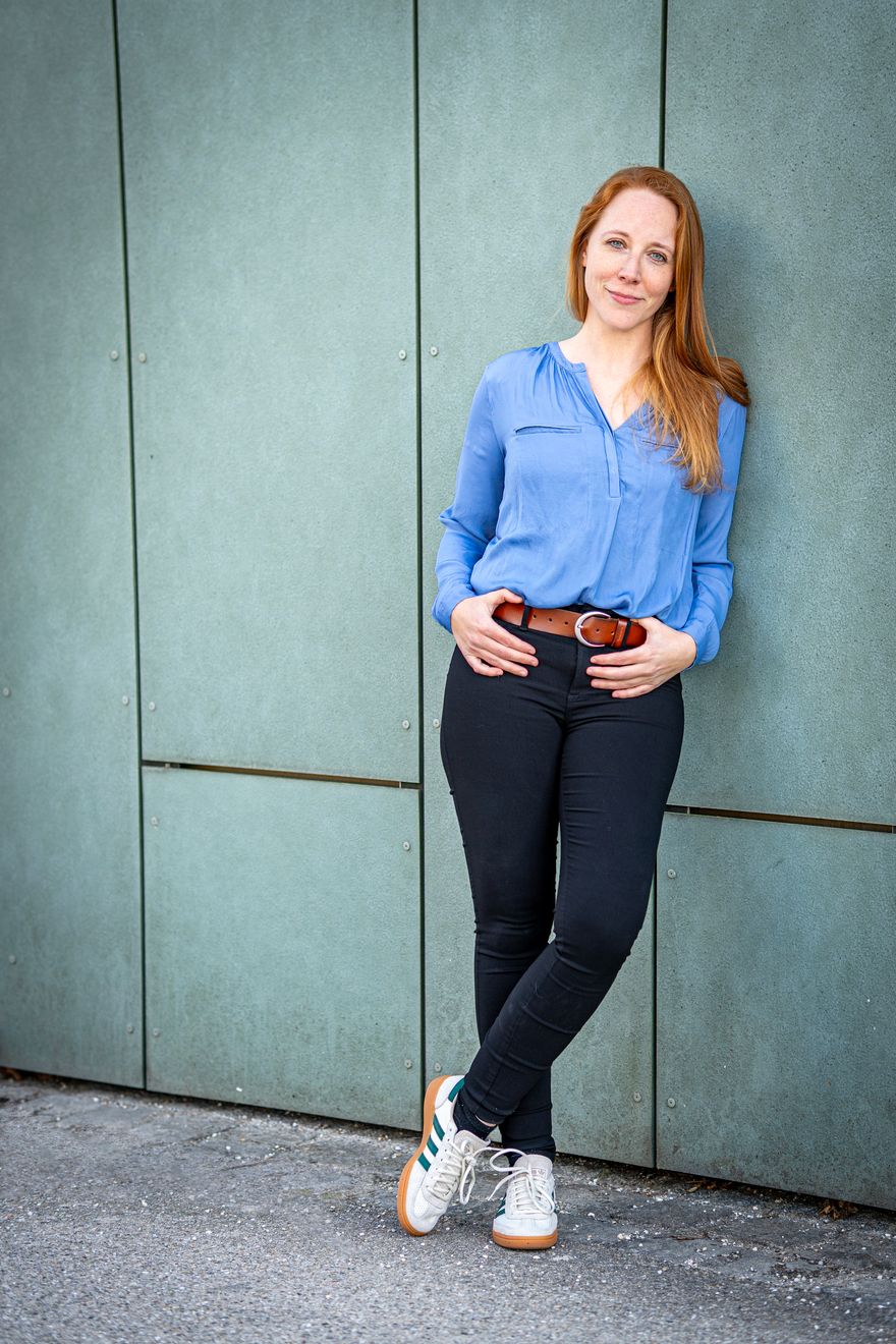 Debora Mergler in blauer Bluse, Jeans und Turnschuhen lehnt entspannt an einer grünen Wand und lächelt in die Kamera